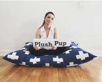 Jona, owner of Plush Pup Inc holding sign that reads the company's name while sitting on a large dog bed